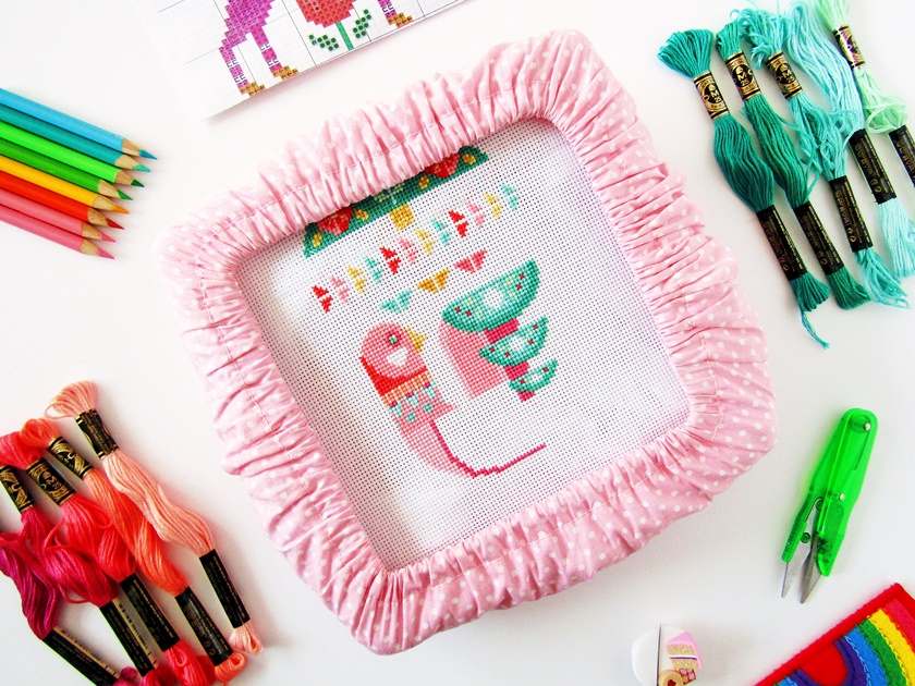 An in-progress photo of a Satsuma Street Deck the Halls Wall Hanging cross stitch, showing a partially completed pink Scandinavian-style bird cross stitch on white aida in a pink polka dot embroidery frame in the centre of the photo, surrounded by a range of pink and teal threads, fluorescent green embroidery scissors, a cake needle minder and needle, coloured pencils, and a cross stitch pattern.