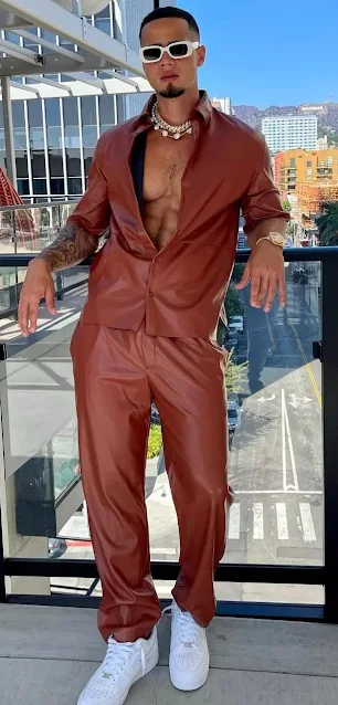 Sexy black dude wearing a red orange burnt sienna leather suit tracksuit outfit looking all pimp sporting large white sunglasses and a big flashy I'm super tacky white Coral chain necklace that's more fashion than ghetto