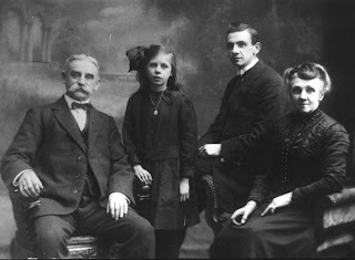 A greyscale photo of four people.  An elderly man with a very bushy mustache, a young girl with a big bow in her hair, a man in his twenties seated on the arm of a chair and in that chair an elderly woman, very smart.  All are wearing dark coloured clothes.