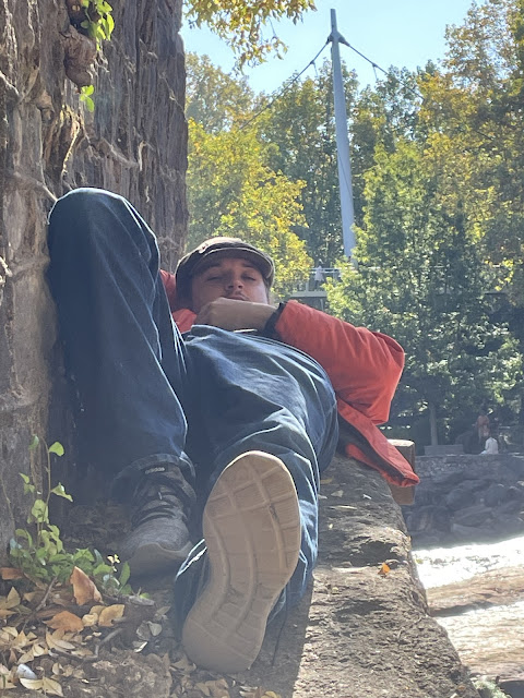 Andy sleeping on a ledge beside the river in Greenville, SC. His right arm is behind his head creating a pillow for himself. He is wearing his sneaker, jeans, his irish cap, and his orange jacket. the river is on his right and a rock wall is beneath him and beside him on the left hand side.
