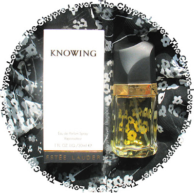 The bottle and the white cardboard box of Knowing by Estee Lauder (eau de parfum) lying flat on a black, airy fabric with white flowers on it