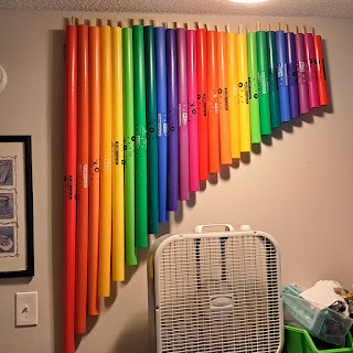 The picture shows some general household items, but the focus is on a chromatic set of Boomwhackers, arranged by color and size, mounted on the wall.