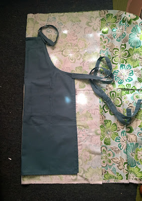 A photo from above of a colonial-blue cooking apron, folded in half vertically and laid on top of a back-folded piece of spashy green, teal, and grey tropical flower print fabric. The folded edges of the apron and fabric are aligned at left, and the apron straps coil and trail across the fabric past the back-turned edge. Bright sunlight dapples everything.