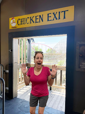 A white woman with dark hair in a pink skirt and navy shorts stands in a doorway with a rollercoaster in the background. Above the doorway is a sign that says "Chicken Exit."