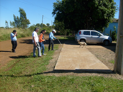 Imagem de uma calçada de concreto, contornada por grama e a rua sem asfalto. Ao fundo tem um carro e um cachorro. Marcos, Ramão, eu e Luiz estamos indo em direção à calçada de concreto.