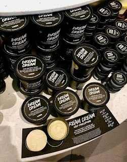 A selection of black cylindrical tubs with dream cream in white font with white cream inside with a rectangular black plaque that says dream cream on a large rectangular light brown shelf on a bright background