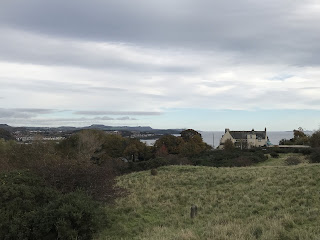 A view over a grassy hill to a white house and on to the coast of Fife.  A memorial stone can been seen sitting in the grass of the hill.  Photo taken by Kevin Nosferatu for the Skulferatu Project.