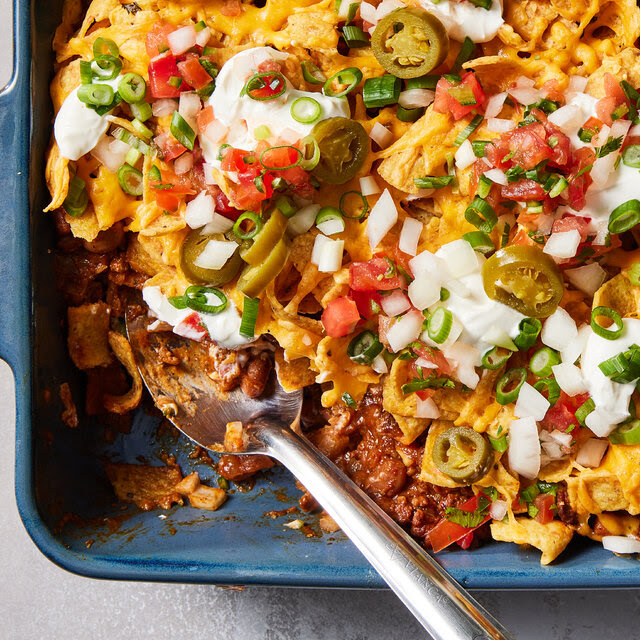 In a dark blue casserole dish, Fritos are layered and mixed up with chili, jalapeños and cheese. The recipe is topped with sour cream, diced onions, pico de gallo and green onions. On the left side, a portion has been taken out with a metal spoon.