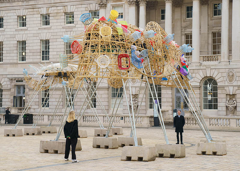 An installation view of The Arks of Gimokudan by Leeroy New. An upturned ship structure made of recycled materials stands in the courtyard, with two people standing either end of the sculpture.