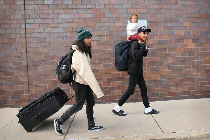 On a sidewalk in front of a brick wall, a woman pulls a suitcase and a man carries a toddler on his shoulders.