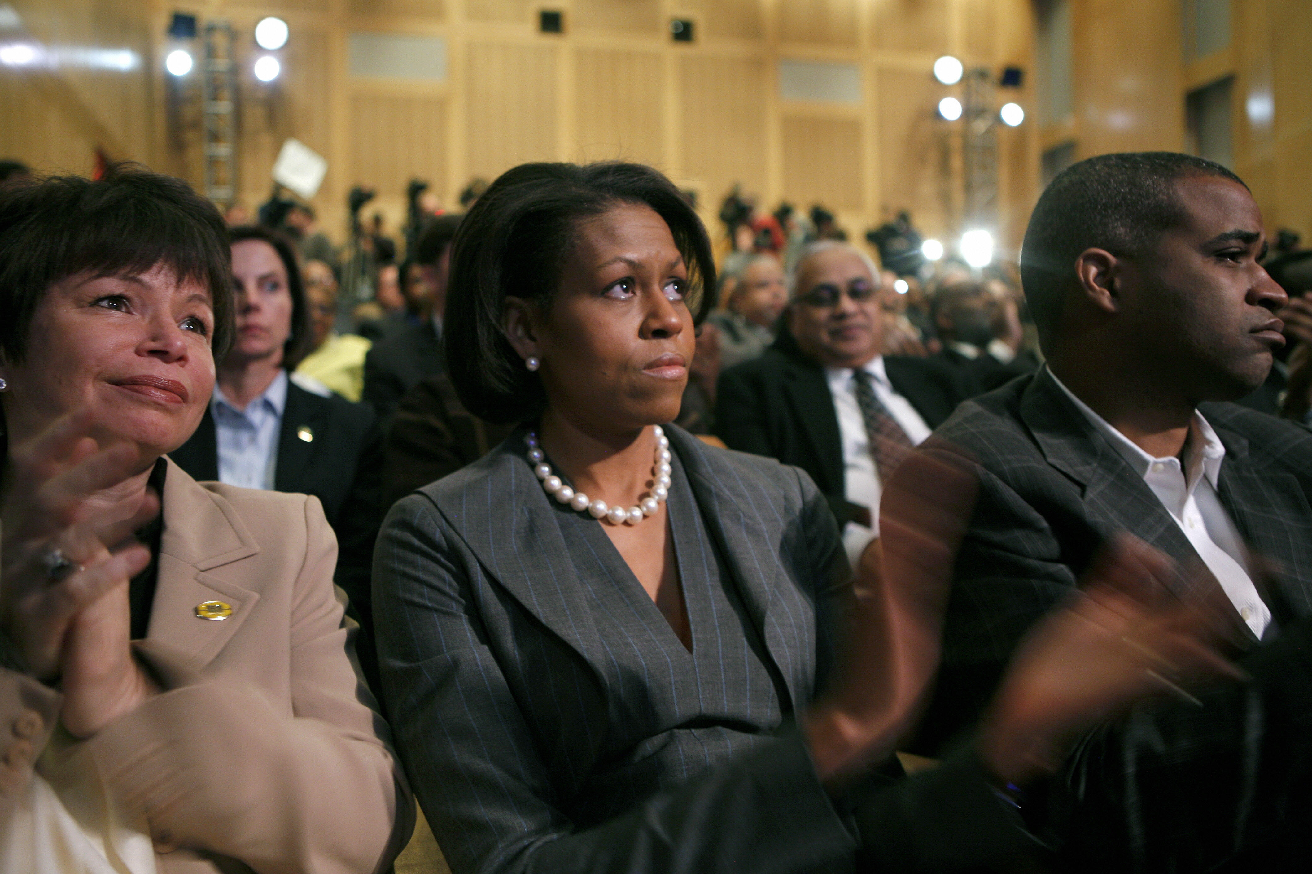 Valerie Jarrett, Michelle Obama, and Marty Nesbitt sit in the audience at the "A More Perfect Union" speech. Valerie Jarrett is wearing a beige blazer and claps as Michelle Obama looks on attentively. Mrs. Obama is wearing a gray jacket and large pearls. 