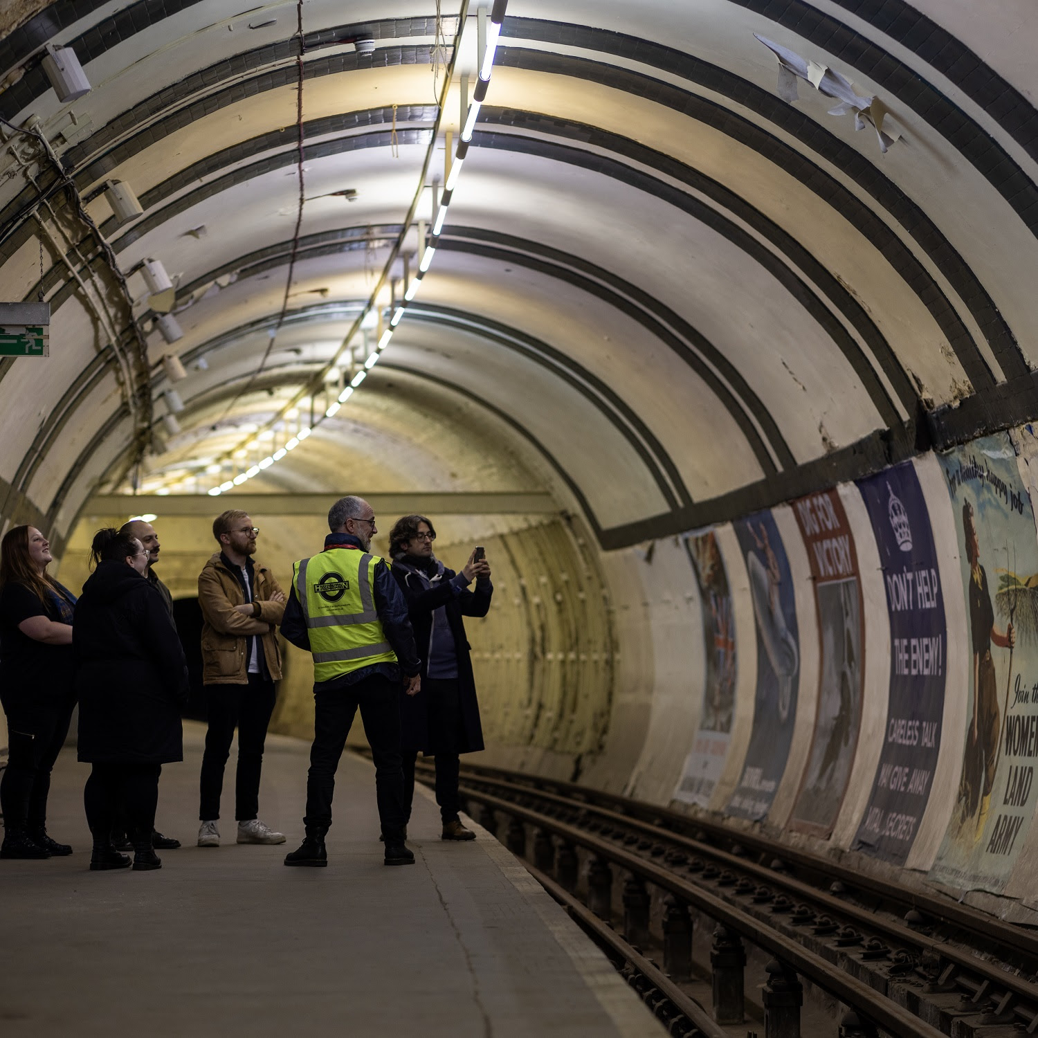 Group of people on Underground station platform taking photos of historic advertising posters