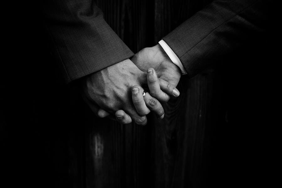 A black and white photograph shows the clasped hands of two people in suit jackets. One has a visible wedding ring.