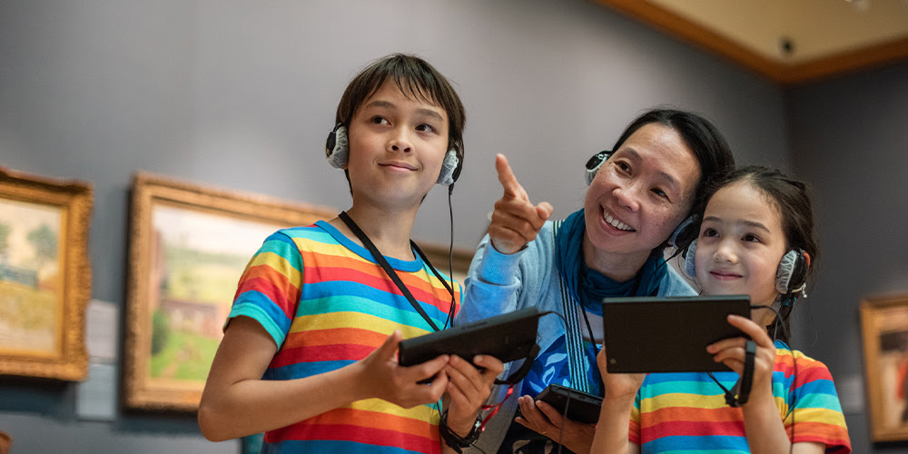 A woman and two children use hand-held multimedia devices and headphones in the Museum