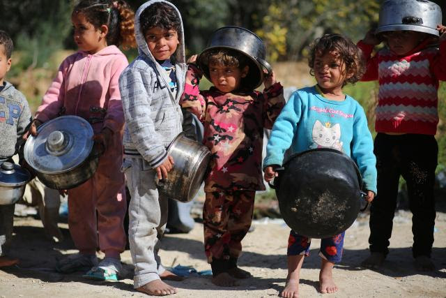 Row of very young Palestinian children holding cooking pots, some wearing the pots on their heads. 