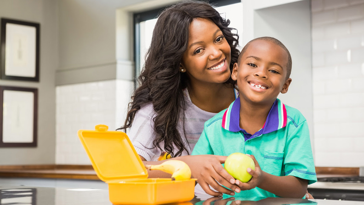 A mother and her young son are holding an apple while smiling at the kitchen counter, where there is an open lunchbox with a banana in front of them