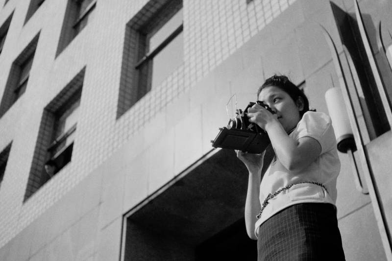 Uma fotografia em preto e branco de uma jovem tirando uma foto com uma câmera.