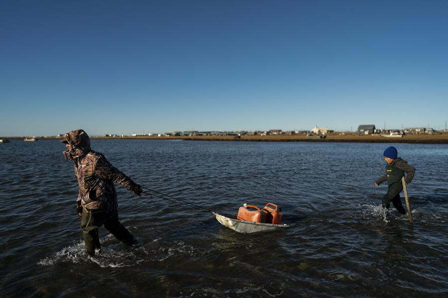 A man walks through water pulling two gas cans on a makeshift float. A child follows behind after him.