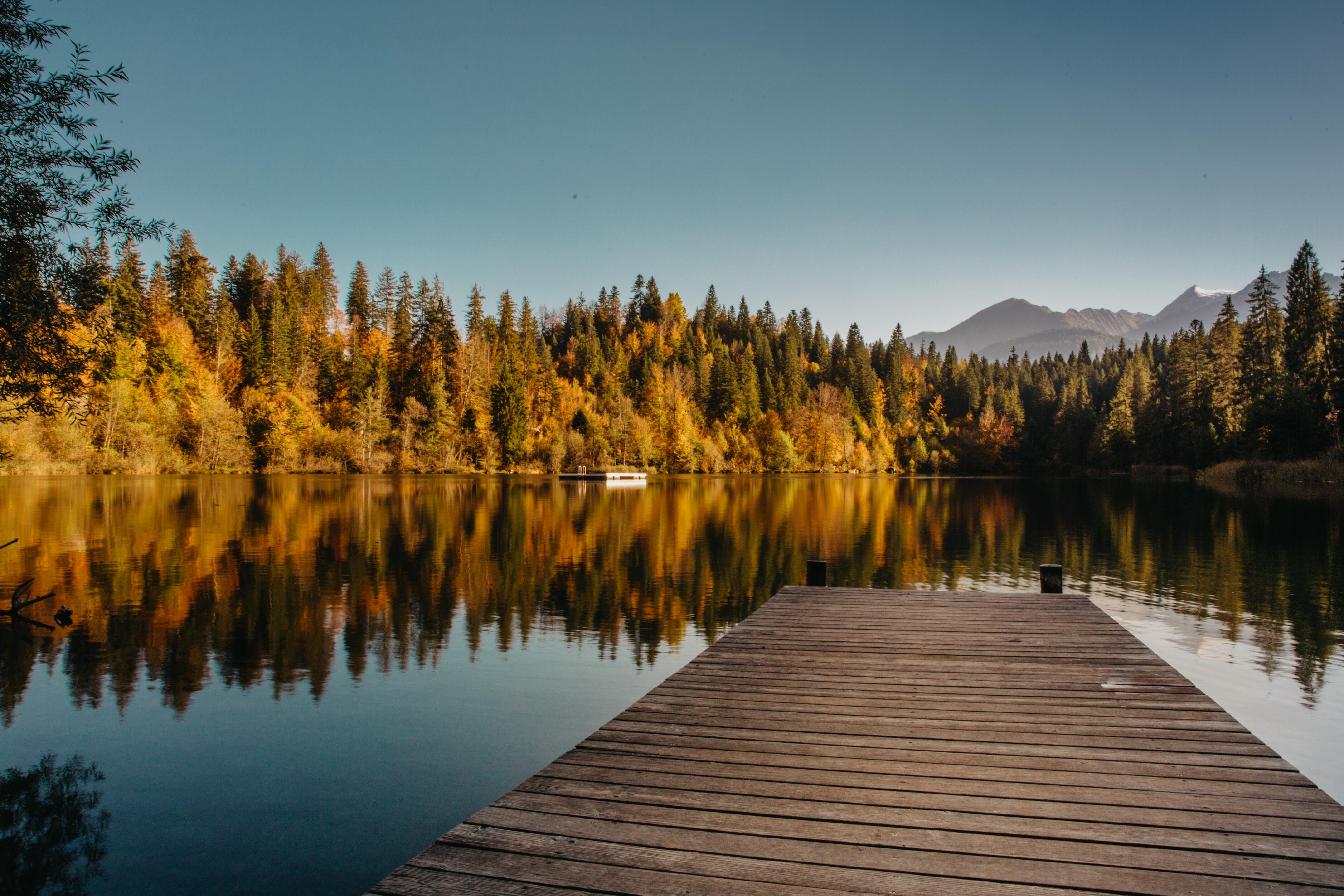 Photo of a dock on a calm lake, surrounded by trees in fall colours, mountains in the background.