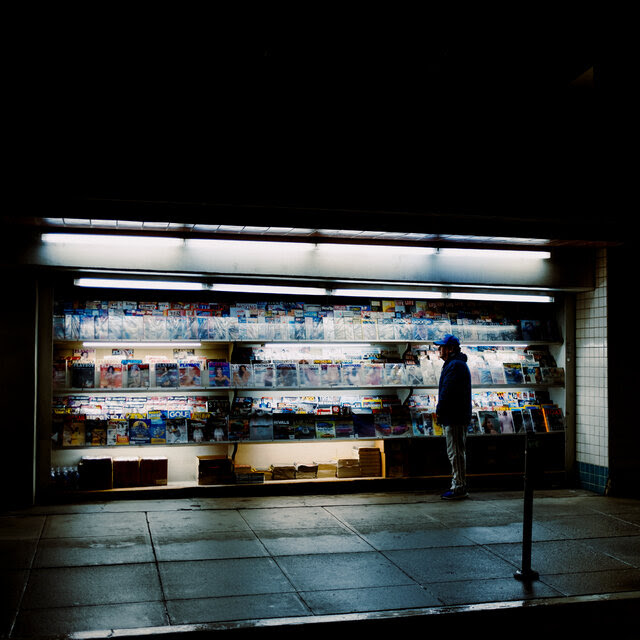A lone figure stands on a sidewalk in front of a newsstand illuminated at night.