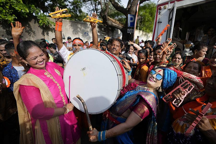 Devotees participate in an annual Rath Yatra, or Chariot procession of Lord Jagannath, in Ahmedabad, India. At the front of the crowd, two people hold a drum between them. Many of the people in the crowd around them have wooden instruments.