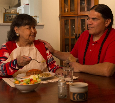 Caregiver rests hand on shoulder of elder eating a meal of salmon, rice, and garden salad.