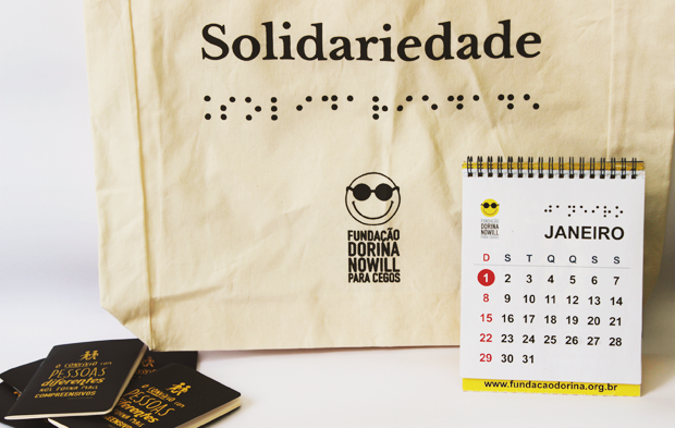 DESCRIÇÃO DA IMAGEM: Foto com fundo branco. A maior parte da imagem é preenchida por uma ecobag de algodão cru com o logo da Fundação Dorina e com a palavra SOLIDARIEDADE em preto e os pontos negros do braille. À frente da ecobag, à direita da imagem, um calendário de mesa mostrando o mês de Janeiro em uma folha branca, com datas em preto e os domingos em vermelho. À esquerda da imagem há uma pilha de caderninhos azuis, com a frase "O convívio com pessoas diferentes nos torna mais compreensivos" em amarelo.