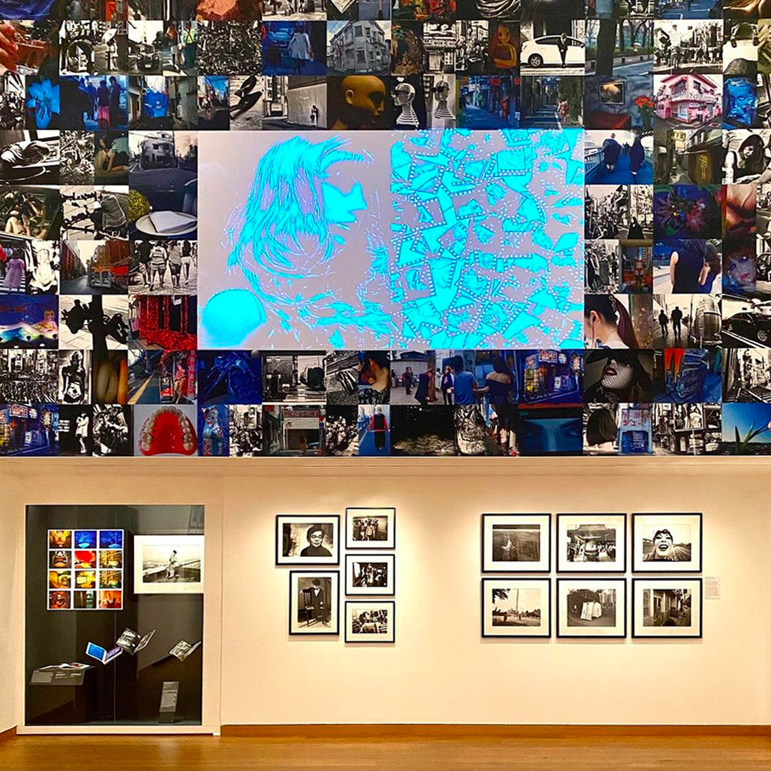 View in the Tokyo exhibition, of framed photographs hung on a wall beneath a large collage of photographs