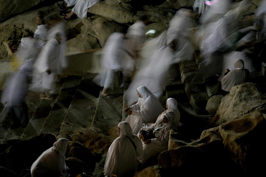 Muslim pilgrims dressed in white clothing pray on a rocky hill. In the background are the blurry figures of other pilgrims walking.