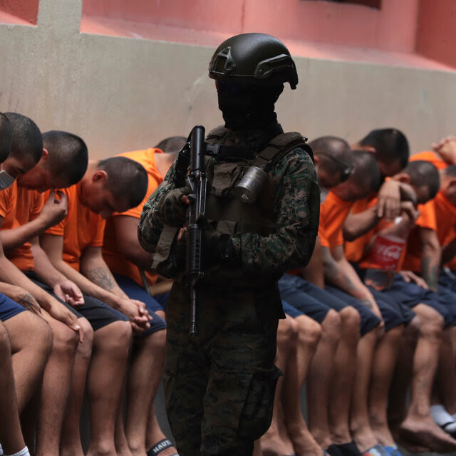 A soldier wearing a face mask, while holding a gun, patrols a line of inmates wearing shorts and T-shirts.