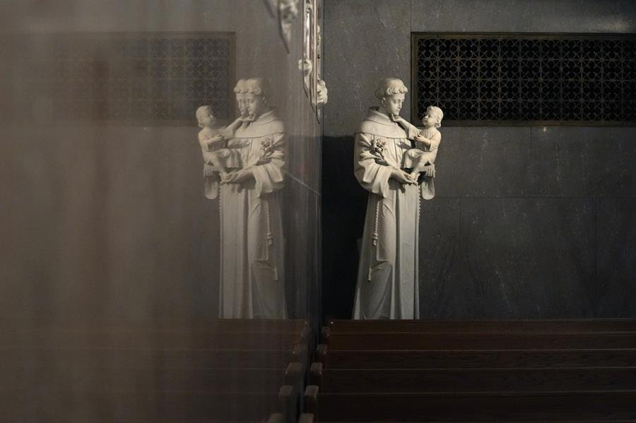 A statue of Saint Anthony holding an infant Jesus. The statue is near a wall that casts a reflection of it.