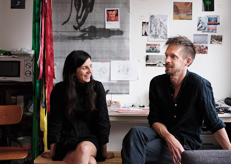 Revital Cohen & Tuur Van Balen sit in their studio. Revital looks at Tuur, while Tuur looks of into the distance.