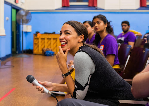 AOC leaning over on a stage with microphone