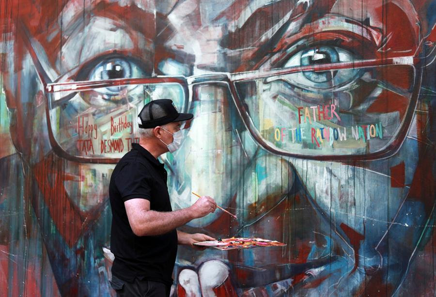 A wall mural of Anglican Archbishop Desmond Tutu is restored by the artist Brian Rolfe. Brian Rolfe stands in front of the mural with a palette in one hand and brush in the other.