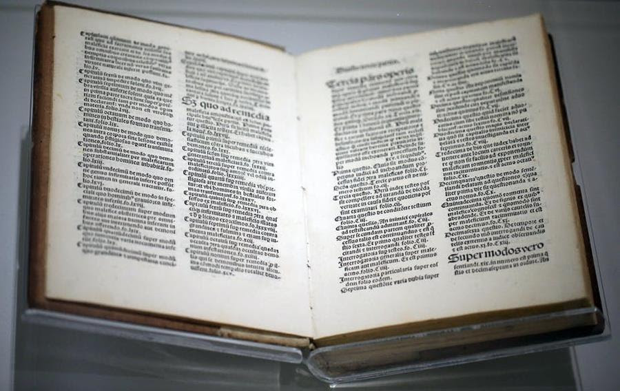 A museum case displays open pages from a medieval manuscript about witches.