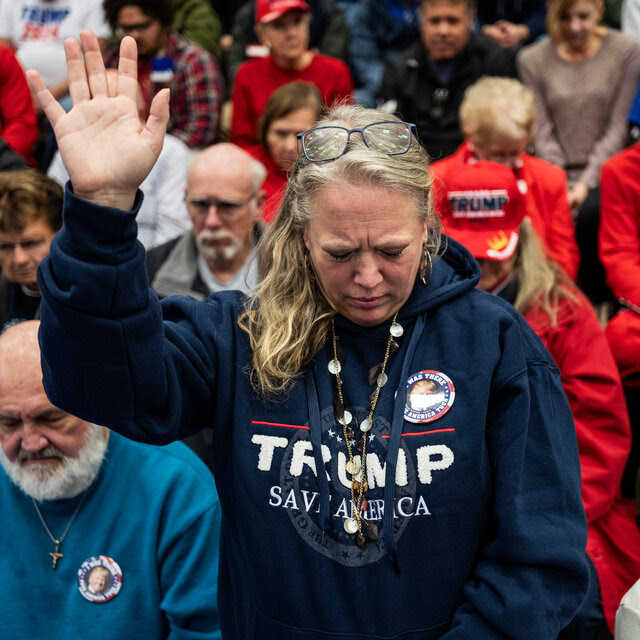 A woman holds her hand up and looks toward the floor as she prays. She is wearing a blue hoodie that reads “Trump Save America.”