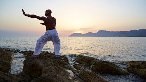  A shirtless man in white pants practices tai chi near a body of water at sunset. 