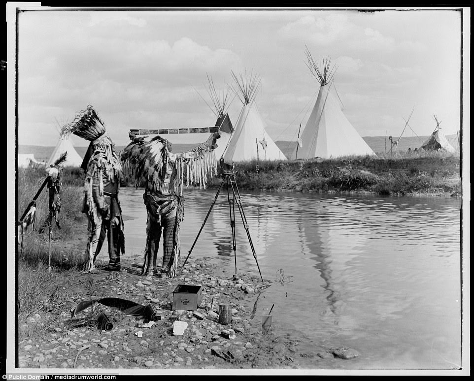 Two Native Americans, wearing feather headdresses, looking at photographic film they stand next to a stream with cameras  at their feet and tipis in the background, in 1913