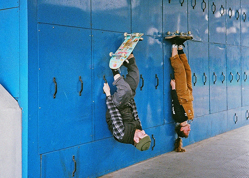 A photo by Dog' Vivaldini of two skateboarders upside down on their boards, against a backdrop of blue lockers.