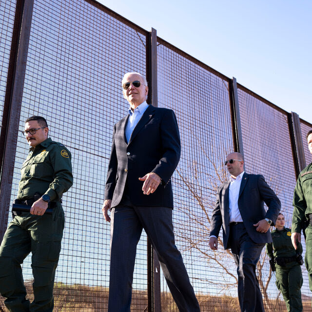 President Biden surrounded by border patrol agents walking near a border fence.