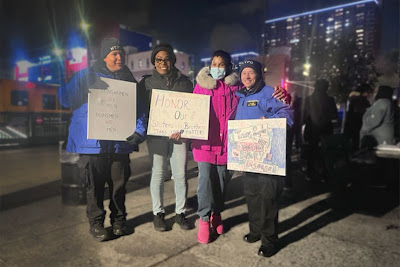Kim Watson-Benjamin smiles on a busy street corner at night time with advocates and NYPD officers. They hold signs with messages like "Honor our Sisters and Brothers."