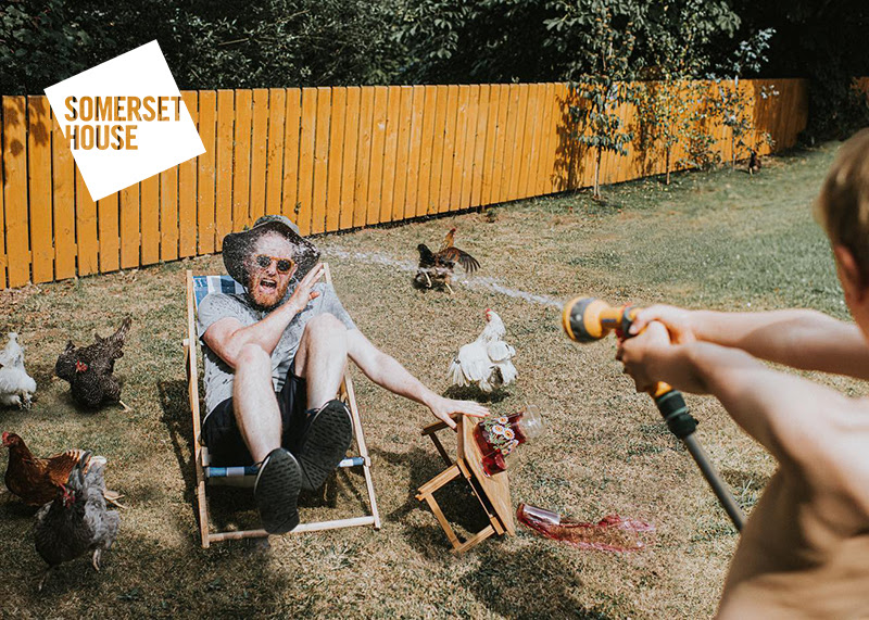 A photo of a young boy using a garden hose to squirt an adult man sat in a deck chair. The image is caught mid-action, with the man opening his mouth in shock as a table next to him is knocked over, drinks spilling everywhere. In the background are chickens who all look to be running away.