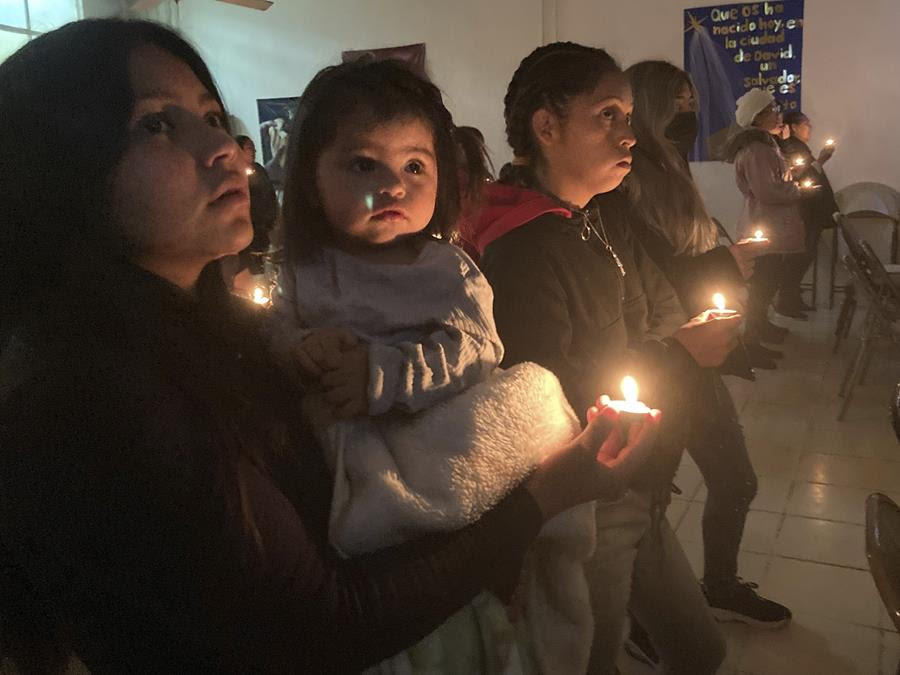 A line of Latinas hold candles that illuminate their faces. The young woman closest to the camera also holds a toddler.