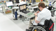 A worker in a wheelchair examines electronics equipment at a desk with a computer and other electronics materials. 