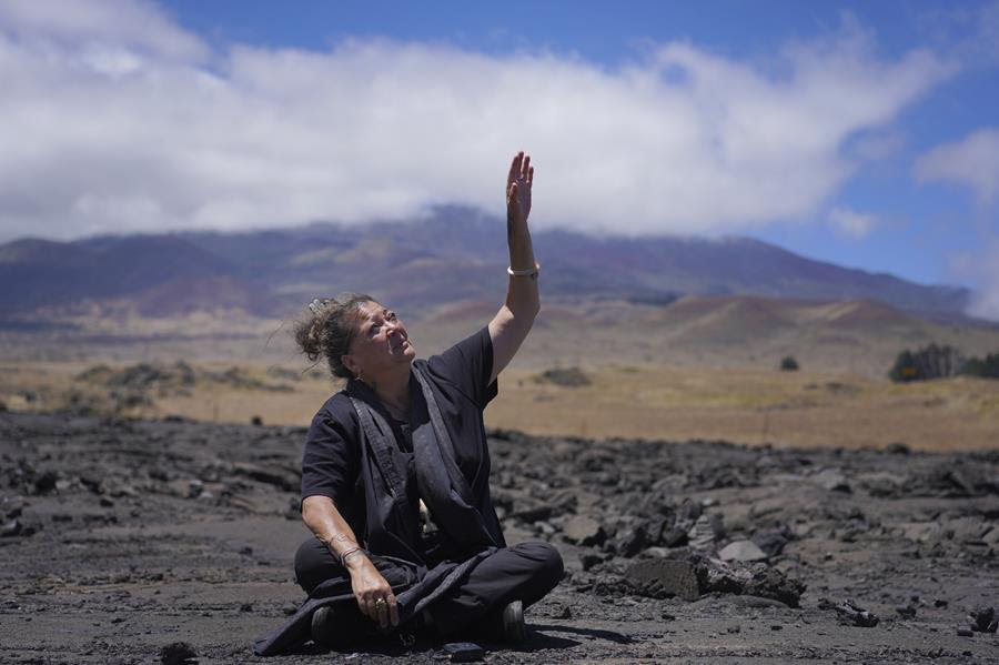 A woman sits on lava rock at Mauna Kea. She has her hand stretched up to the sky.