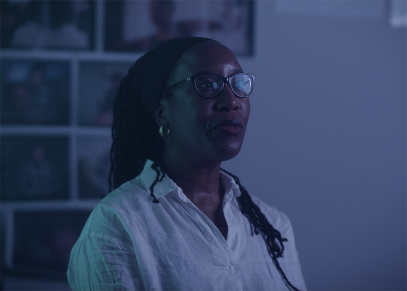 Sonya Dyer on set while filming her artist profile. Sonya looks to a point off camera, and in the background are neatly placed photographs on a wall.