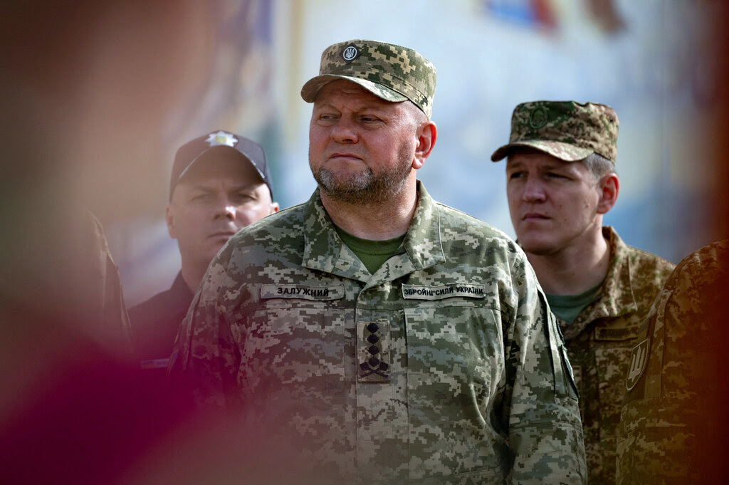 A general in a cap and camouflage stands with other military figures.