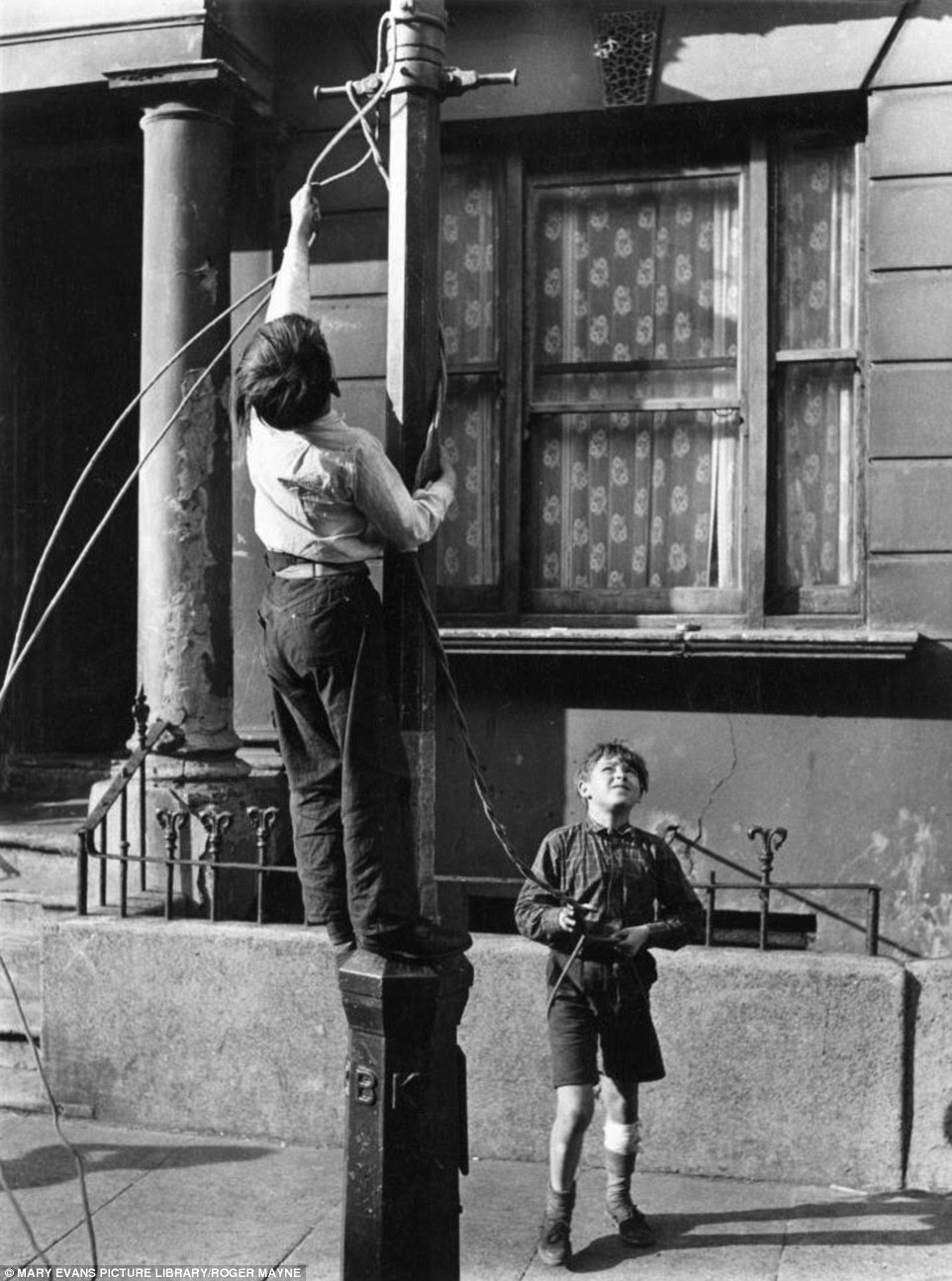 A boy clambers up a lamppost to tie some rope so he and his friend can swing off it in Kensington in 1956