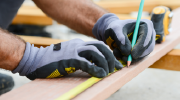 Close-up shot of a worker’s gloved hands marking a wooden plank with a pencil above an extended measuring tape. 