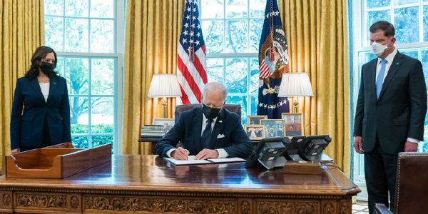 President Biden signs a document in the Oval Office while Vice President Harris and Secretary Walsh look on.